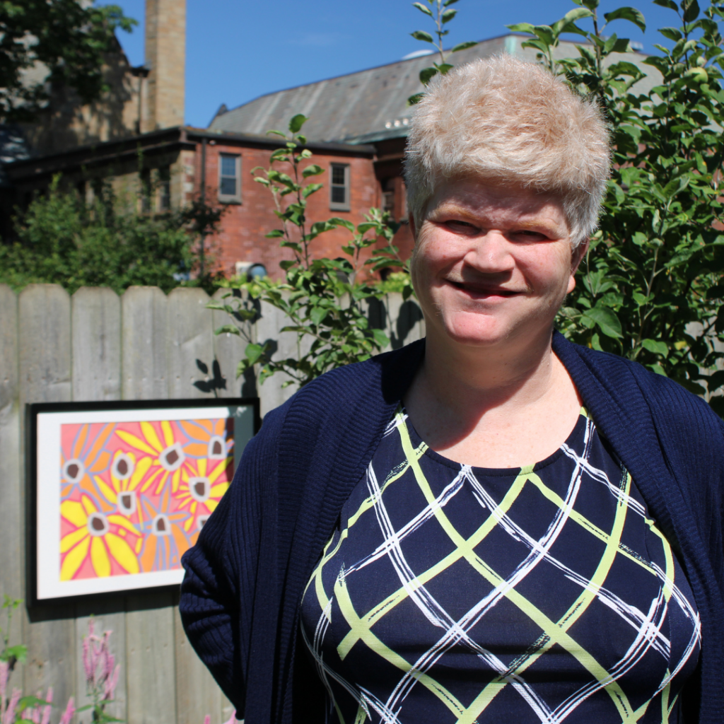 Stockade Art Show 1 Alt Text: A smiling woman with short blonde hair standing outdoors in front of a wooden fence and leafy green foliage. She is wearing a dark blue cardigan over a patterned top with yellow, white, and navy geometric crosshatch designs. To her left, framed artwork featuring colorful yellow flowers on a pink background hangs on the wooden fence, with a brick building visible in the background under a clear sky.